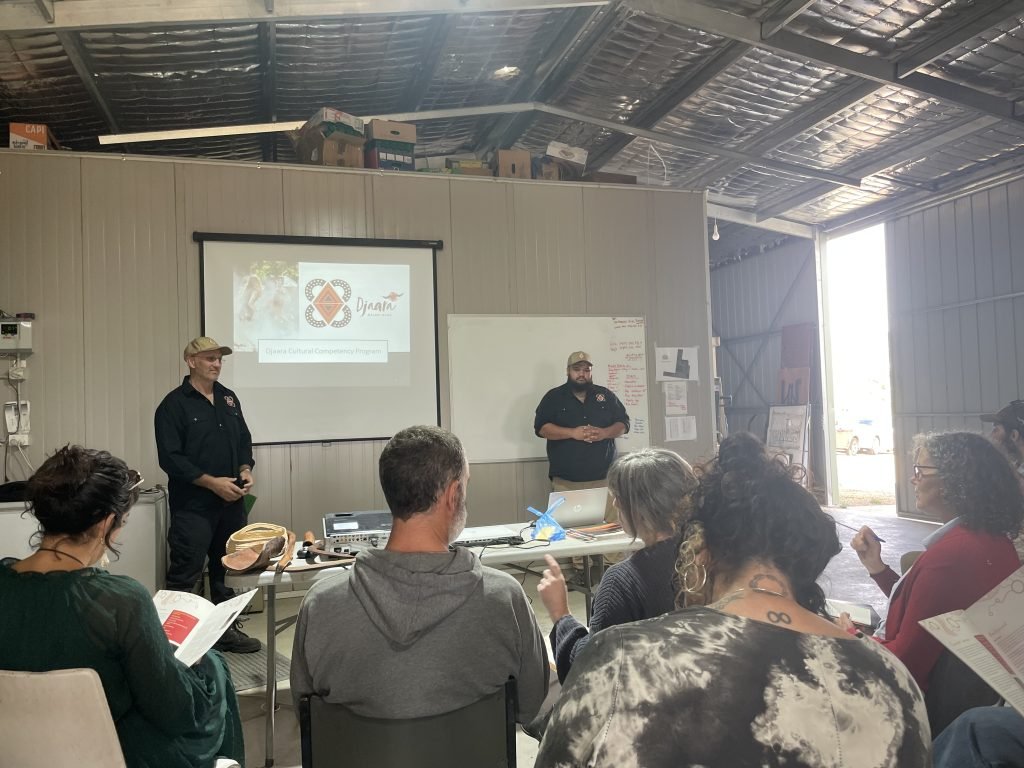 A group of people are sitting in the fruit packing shed at Harcourt, looking at a presentation and two presenters from Djaara