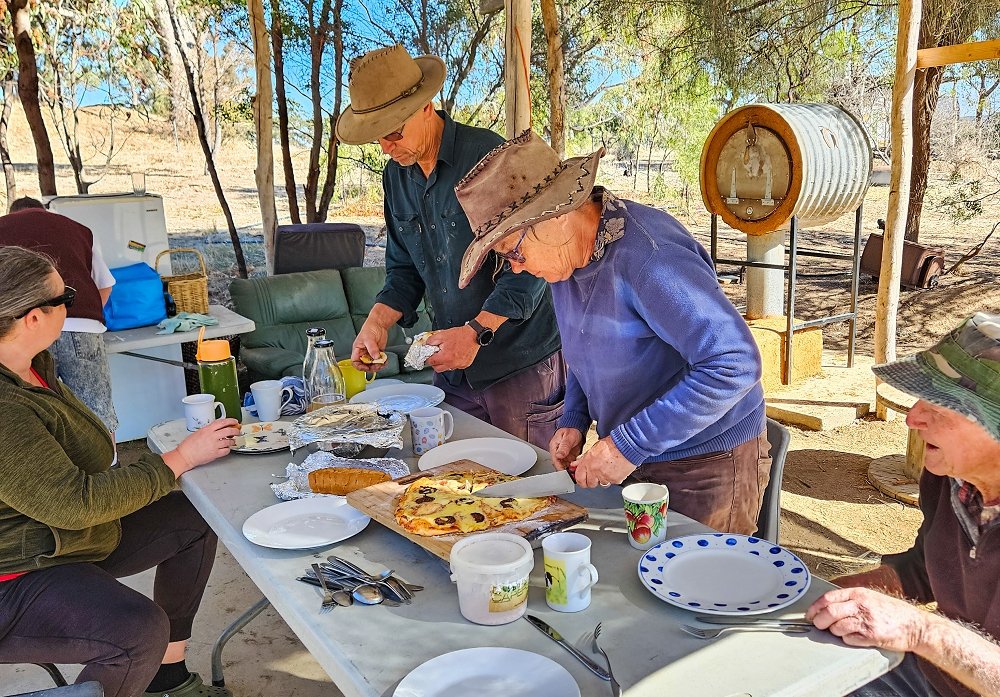 A group of people bending at a table sharing pizza. A woman in a leather hat and blue jumper is cutting the pizza. In the background is a round metal rocket oven on a pedestal with a cob base.